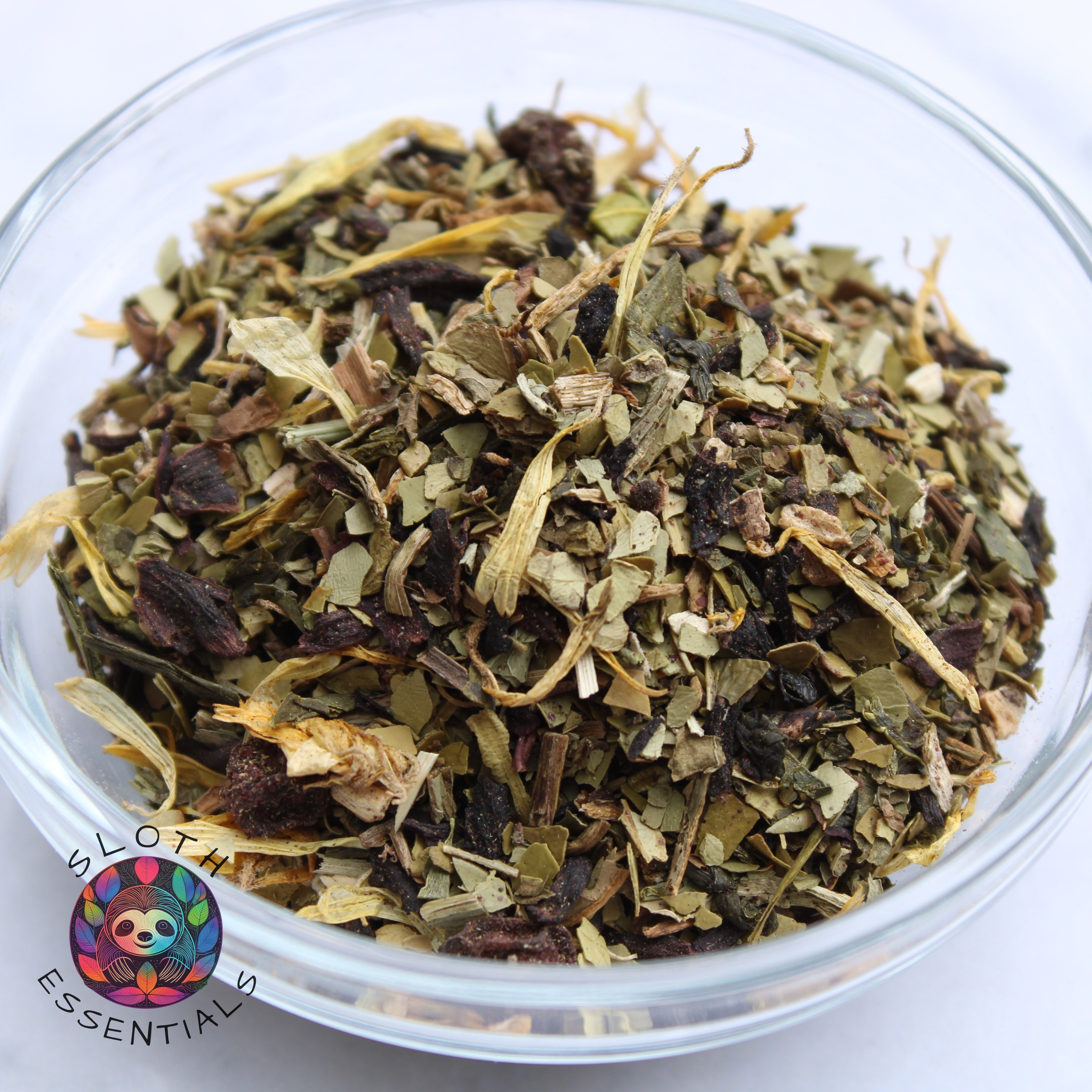 Close-up of Sloth Slim Down Tea ingredients in a glass bowl, showcasing burdock root, yerba mate, dandelion leaf, hibiscus flowers, green tea, acai berry, schisandra berry, garcinia cambogia, and calendula petals.